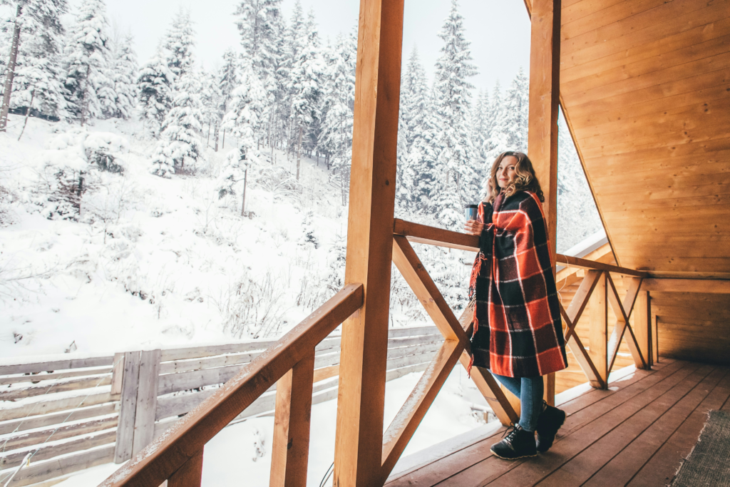 A women, wrapped in a blanket, stands on a porch overlooking a snowy forrest