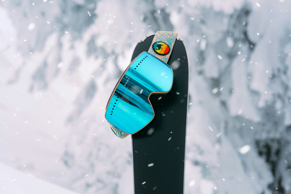 blue ski goggles hang from a black ski as snow floats down from the sky