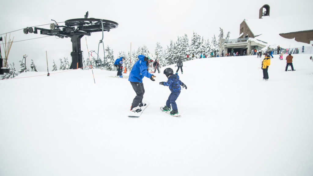 a ski instructor teaches a child how to snowboard at the top of a busy mountain