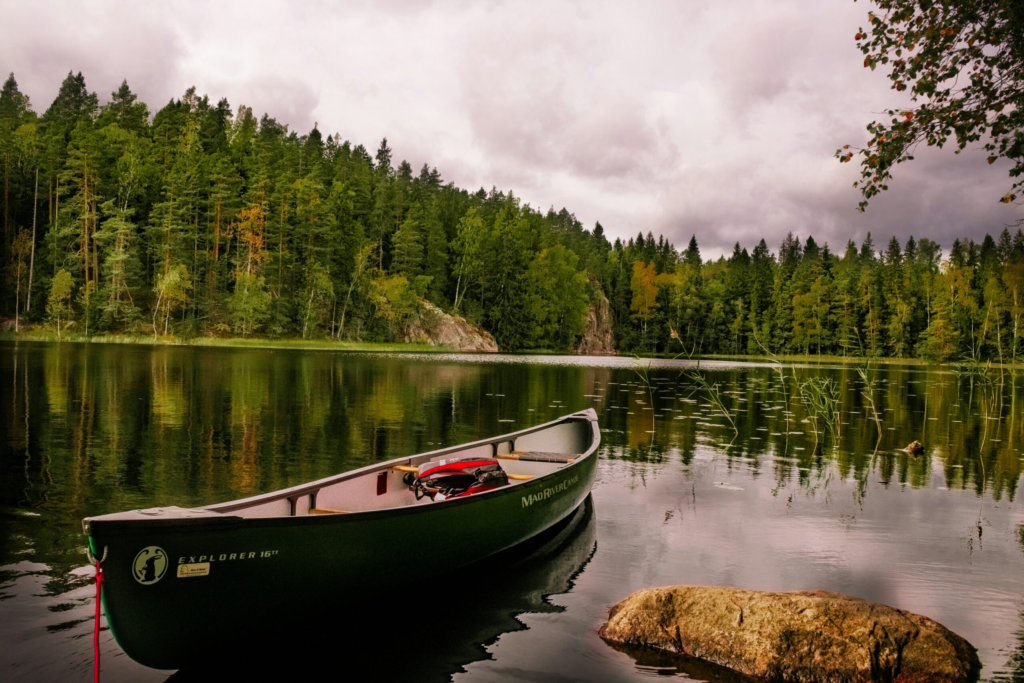 a green canoe tied to the shore sits on a still glassy lake surrounded by green trees