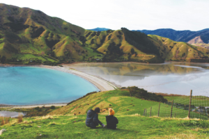 two people on a working holiday visa sit on a grass hill and enjoy the view of the New Zealand mountains and lake
