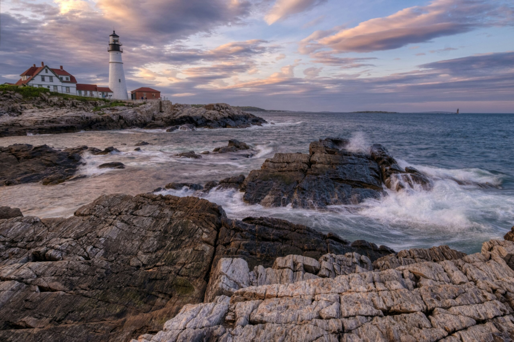 a rocky beach in New England with waves crashing against the shore and a white and red light house in the distance