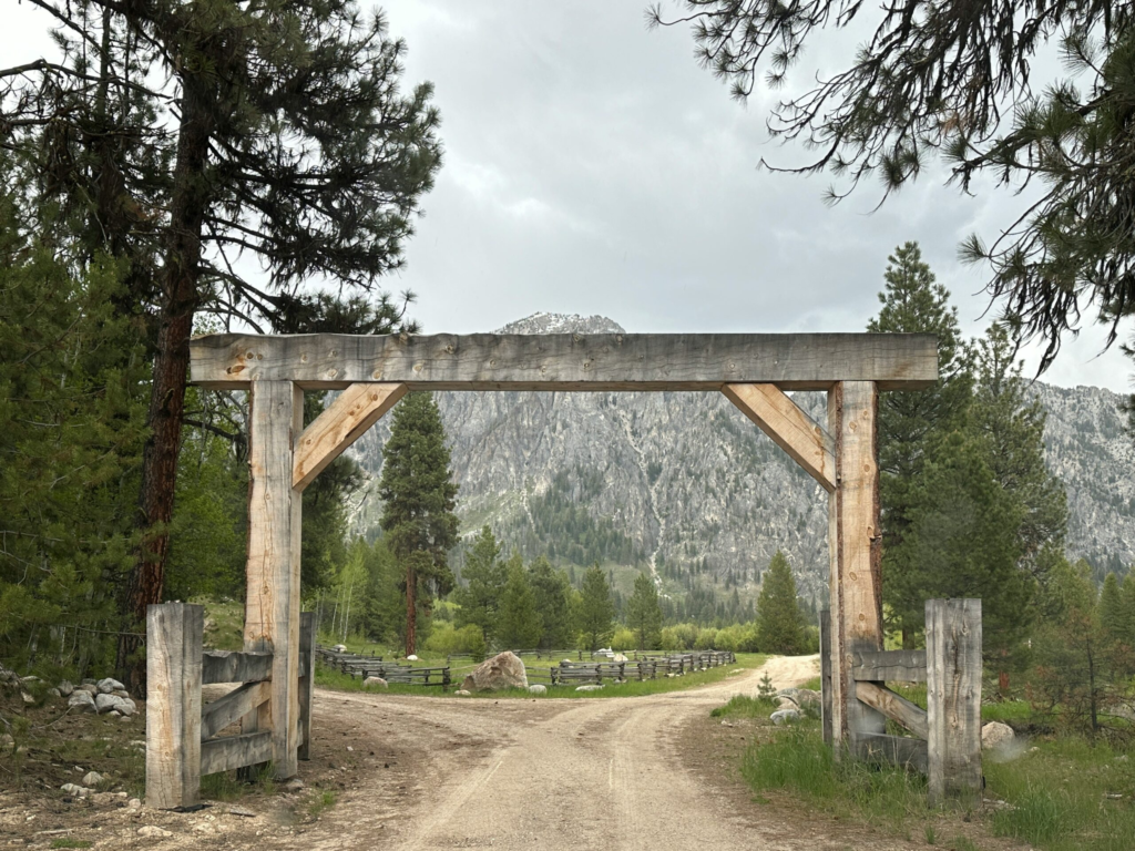 a wooden arch opens to a dirt road, the entrance to a guest ranch in the mountains