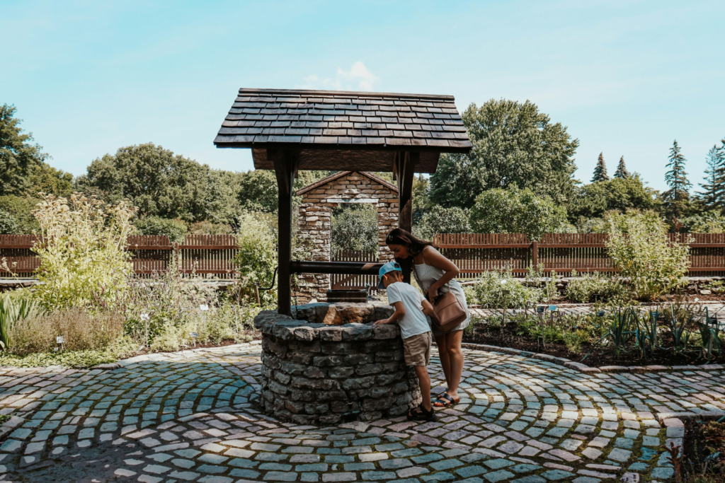 An Au Pair and her host child look into a wishing well while exploring a garden in the summer time